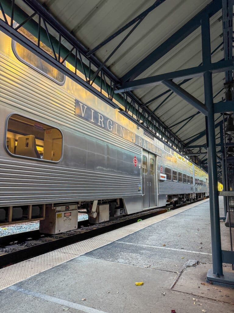 A silver Virginia Railway Express train is stopped at an outdoor station platform under a metal canopy, with empty tracks and scattered debris on the concrete platform.