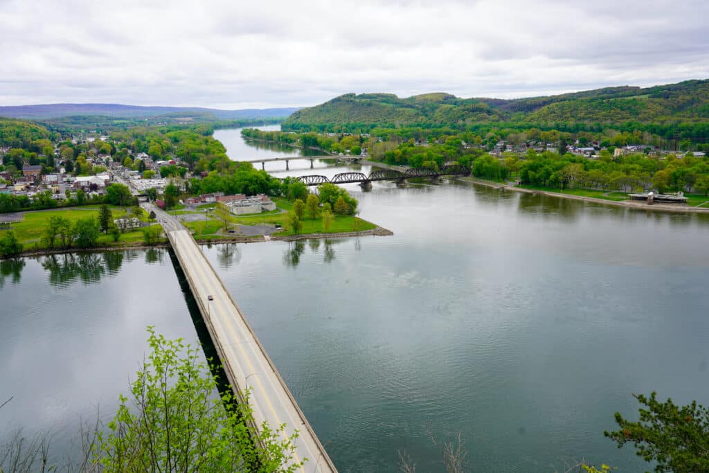 A wide river in Susquehanna River Valley with two bridges crosses through a green, hilly landscape. A small town is visible along the riverbanks under a cloudy sky, with roads and trees surrounding the water.