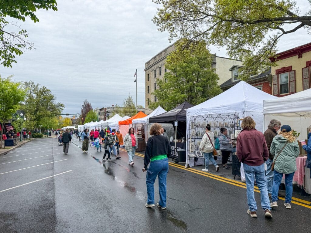 People walk along a street lined with white and colorful vendor tents at an outdoor market in the Susquehanna River Valley. Some browse stalls with art and goods, while others stroll or chat. Trees and buildings border the street.