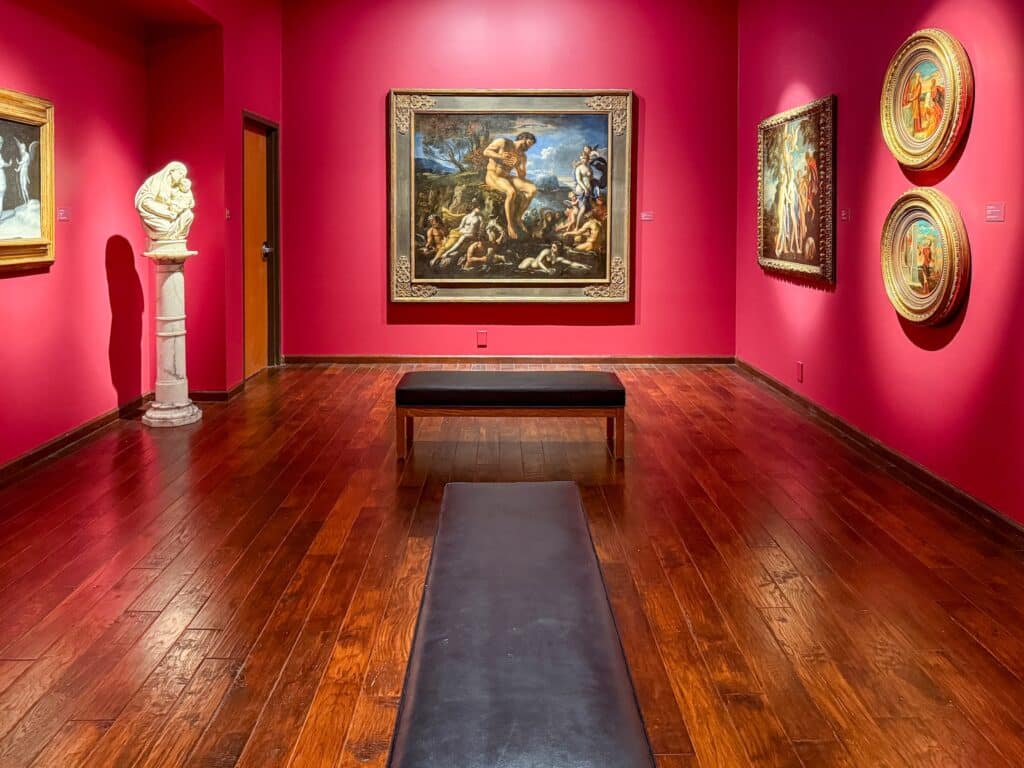 A brightly lit art gallery with vivid red walls, wooden floors, two benches, classical paintings—some depicting the Susquehanna River Valley—in ornate frames, and a white marble sculpture on a pedestal.