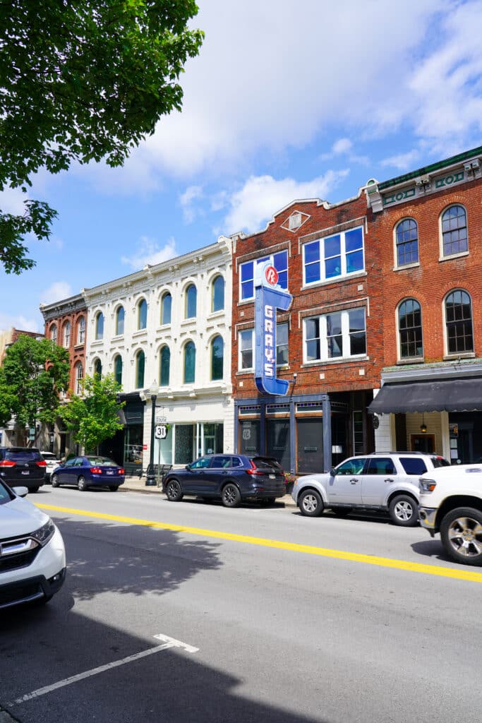 A city street with parked cars sits in front of historic brick and white buildings—perfect for those seeking things to do in Franklin TN. A vertical GRAYS sign adorns one building, while trees and a blue sky with clouds complete the scene.