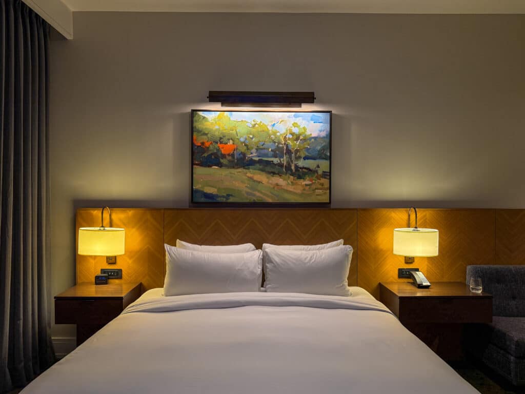A neatly made Harpeth Hotel bed with white linens is flanked by two nightstands with lamps. Above the bed hangs a colorful landscape painting, while guests can unwind before exploring things to do in Franklin TN, seen through the gray curtain and chair to the side.