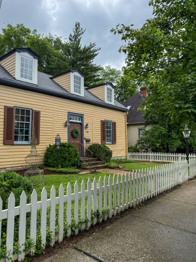 A yellow house with brown shutters, white trim, and a wreath on the door sits behind a white picket fence—capturing the charming vibe you’ll find among things to do in Franklin TN. Green bushes and trees surround it, and the sidewalk is wet from rain.